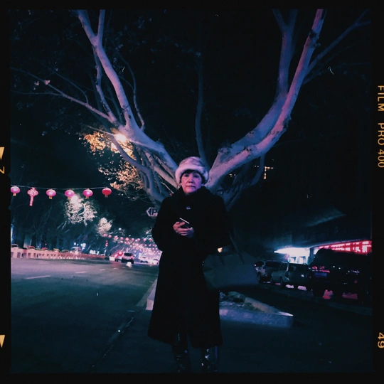 一位穿着外套的女人站在夜晚点亮的树下人行道上。