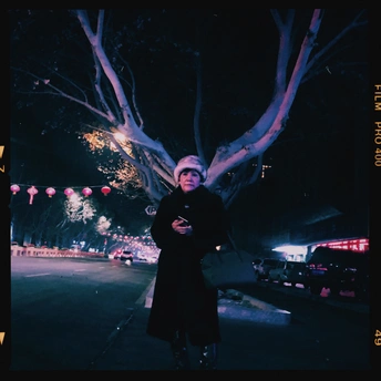 一位穿着外套的女人站在夜晚点亮的树下人行道上。