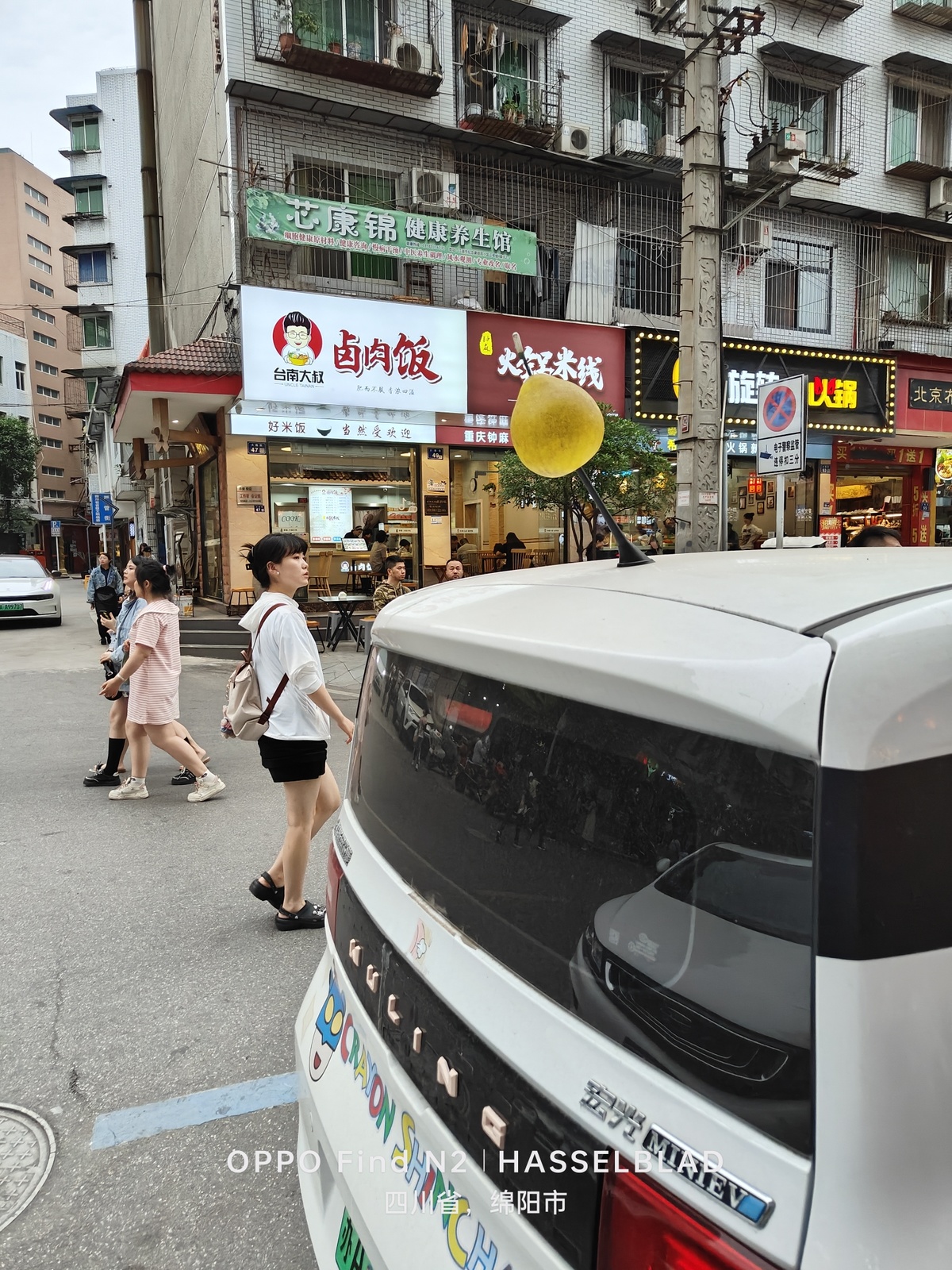 一条人行道上有行人行走 路中央有一辆带有黄色气球的白色汽车。
