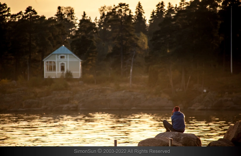 夕阳下 一位坐在岩石上的男子 前方是小型湖泊 背景中有一座白色房子。