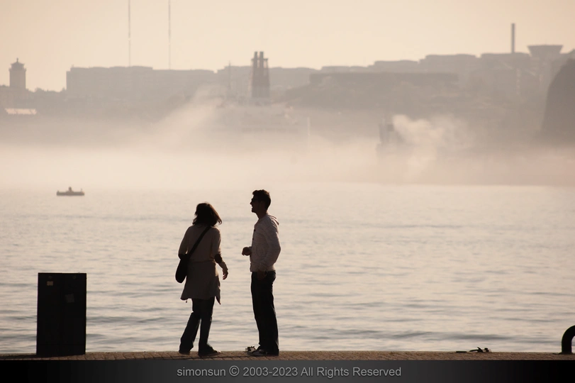 一个人和一个女人站在一个水体旁边 背景是一个城市。