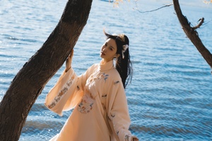 一位穿着外套和裙子的女孩站在靠近水边的一棵树旁
