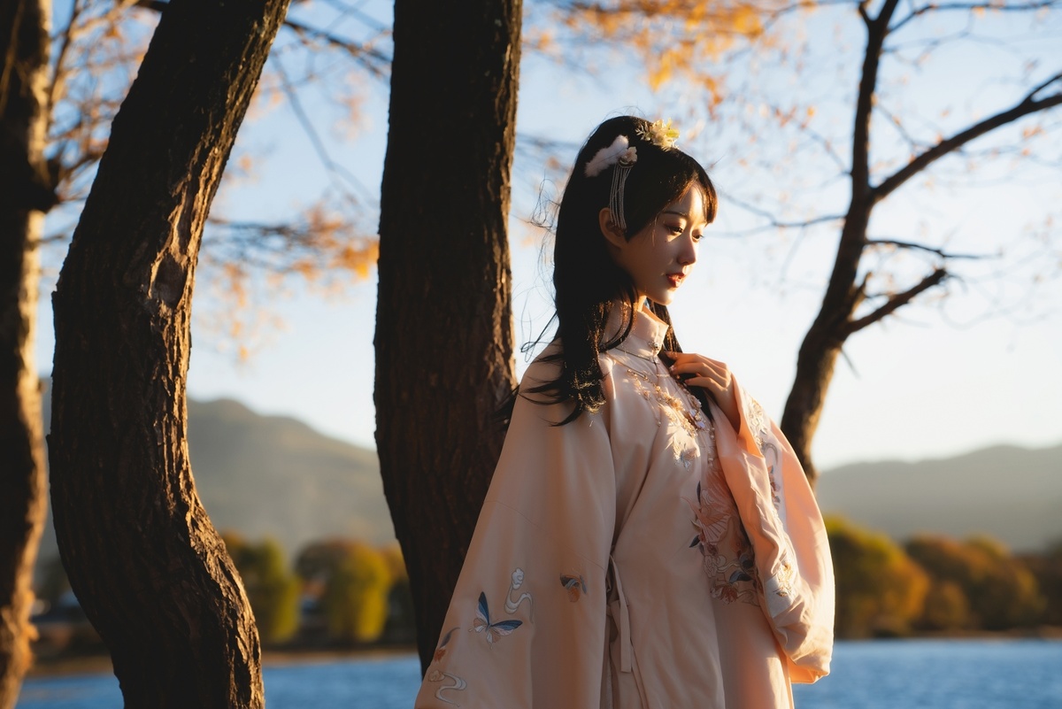 一位穿着和服和外套的年轻女子站在树木和水边。