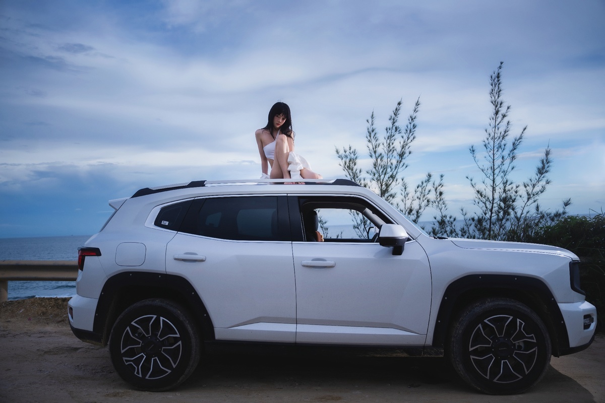 一个年轻女子坐在海滩上的一辆白色吉普车顶部 而一名女子站在车辆后面。