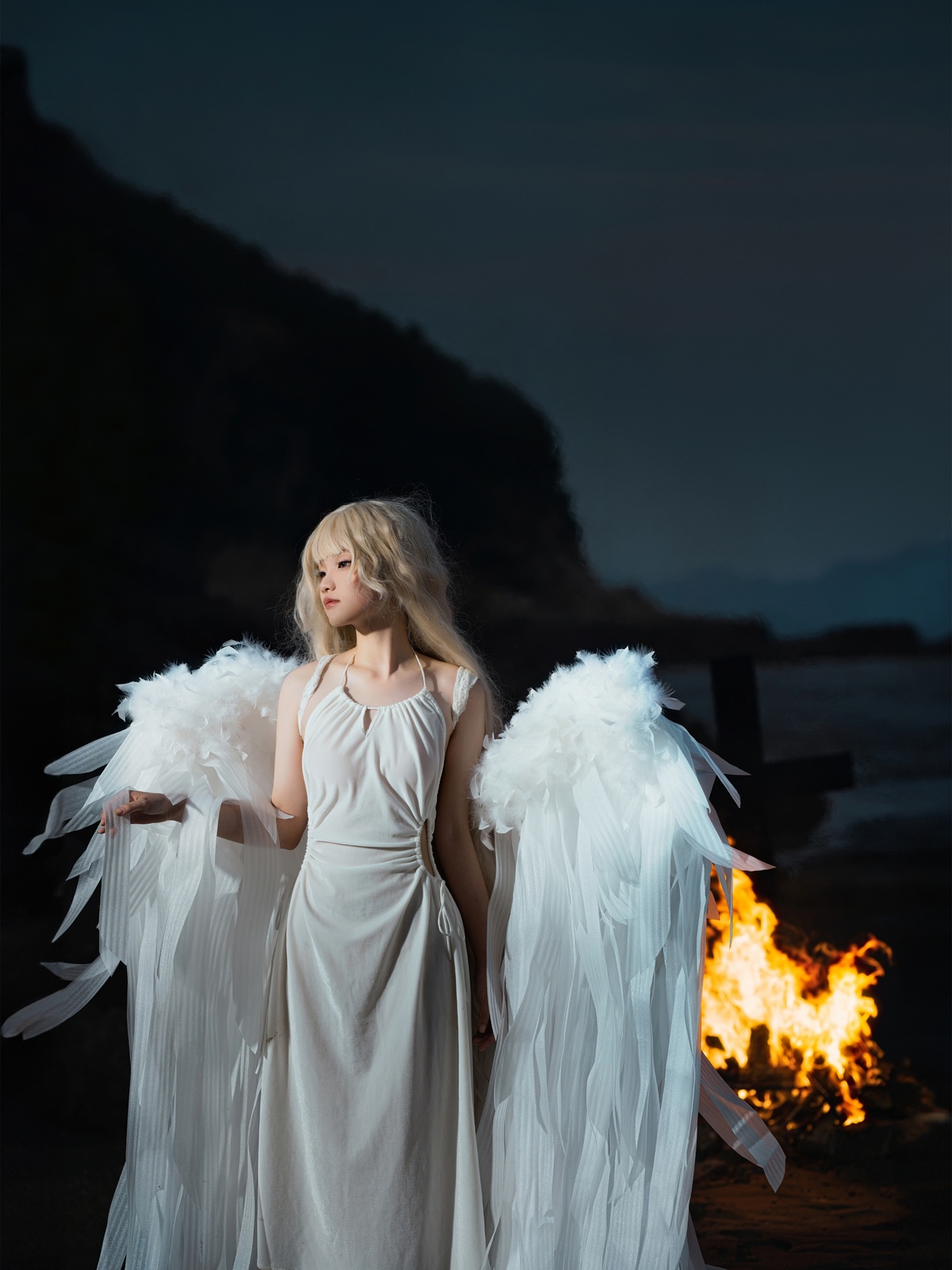 一个穿着天使装 带着白色羽翼的女性站在火前。