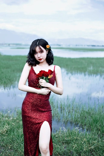 一位身穿红色连衣裙、手持玫瑰的美丽年轻女子站在一片水域旁的田野里