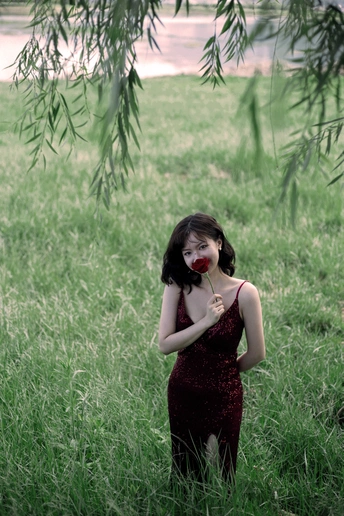一个穿着红色连衣裙的年轻女子站在高高的草地上 手里拿着一朵花