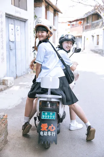 一个穿着制服的年轻男子和女子骑着滑板车沿着街道向下行驶 一个年轻女孩坐在摩托车上。