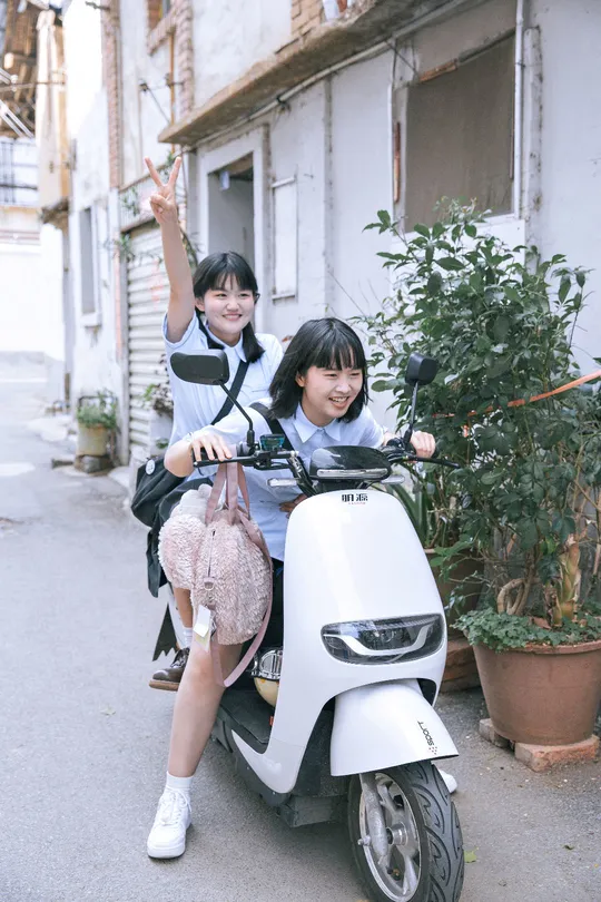 一些年轻人骑着摩托车在一条街道上 旁边有一栋建筑 一个女孩坐在摩托车上。