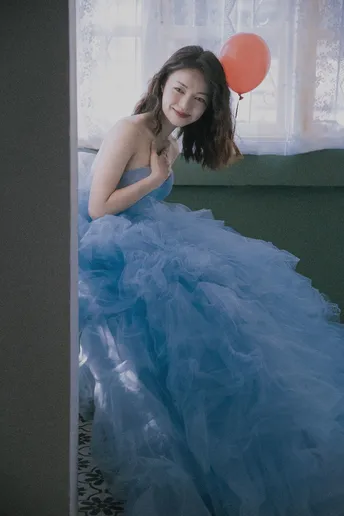 一位穿着蓝色连衣裙的年轻女子在窗户前摆姿势 手里拿着一个红气球。