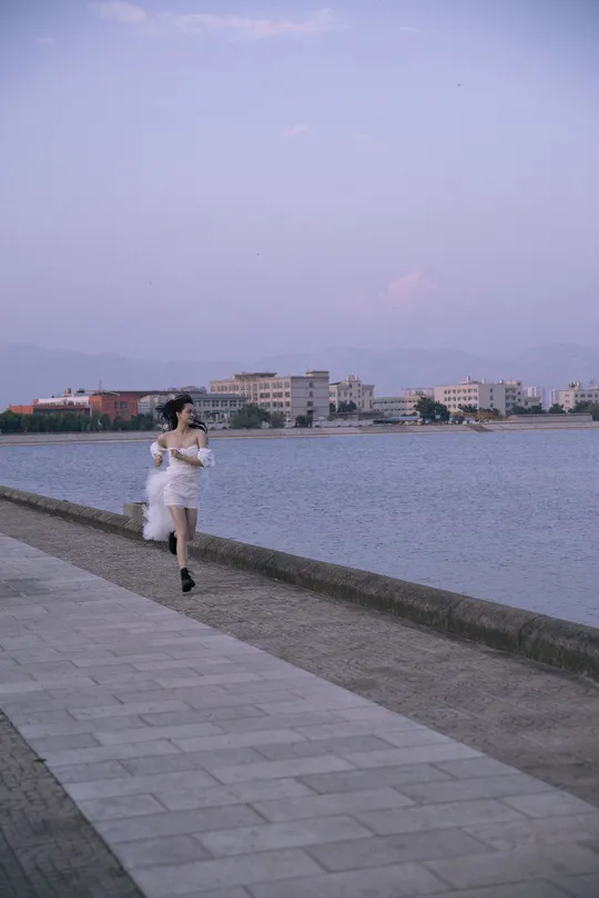 一位穿着白色连衣裙的女子沿着靠近水域的步行道奔跑 背景是湖泊。