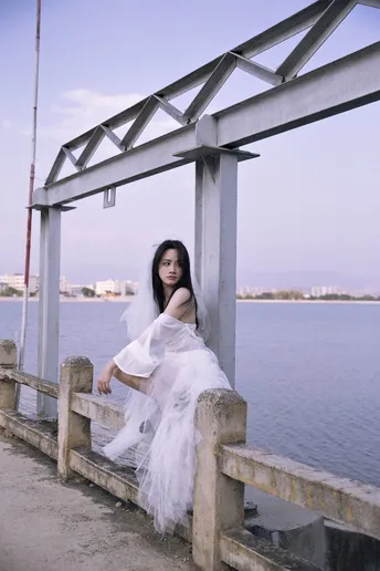 一个穿着白色连衣裙的女人坐在码头上 眺望着一片水域。