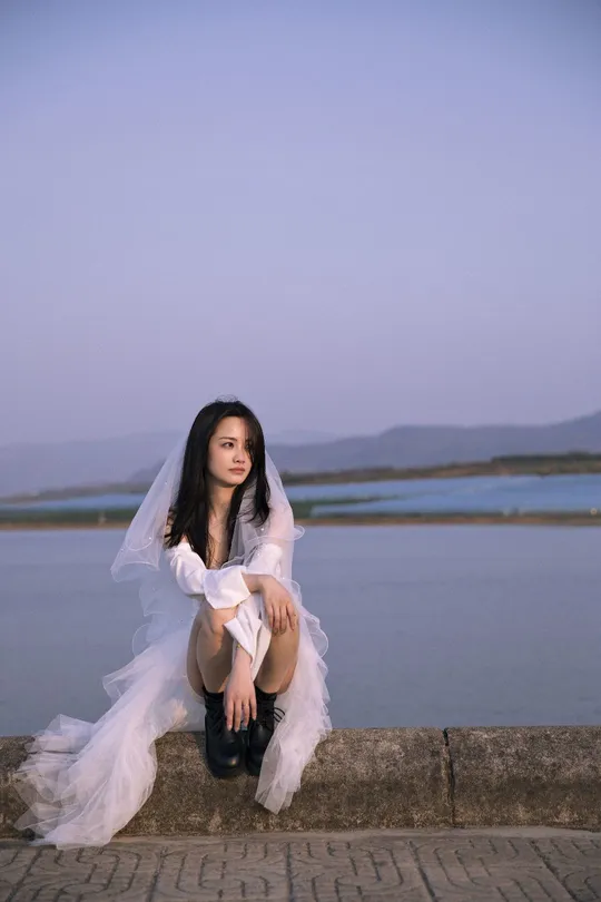 一位美丽的年轻女子穿着白色连衣裙坐在俯瞰一片水域的墙上。