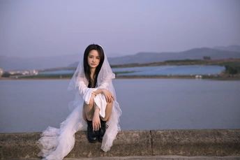 一位美丽的年轻女子穿着白色连衣裙坐在俯瞰一片水域的墙上。