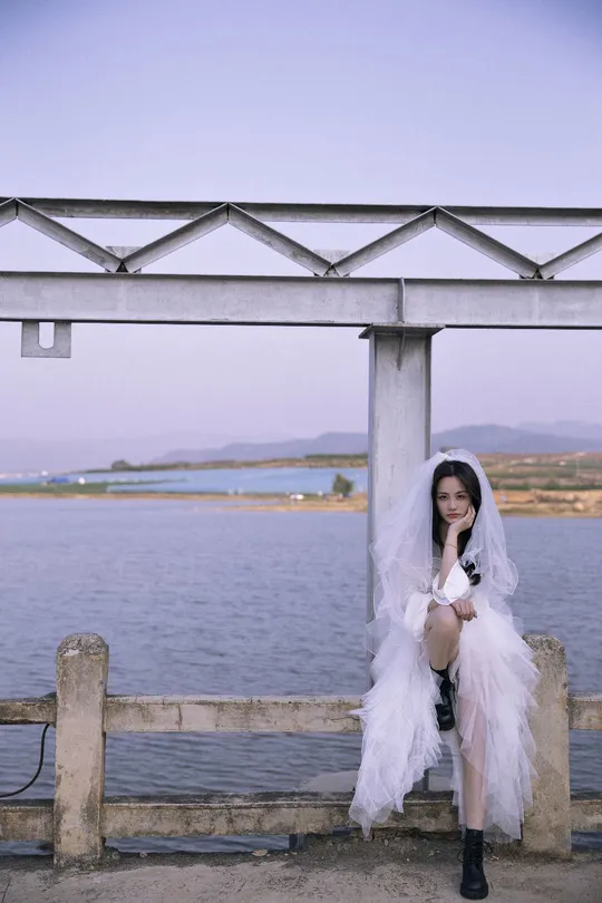 一位穿着白色婚纱的女人坐在码头上 眺望着一片水域。