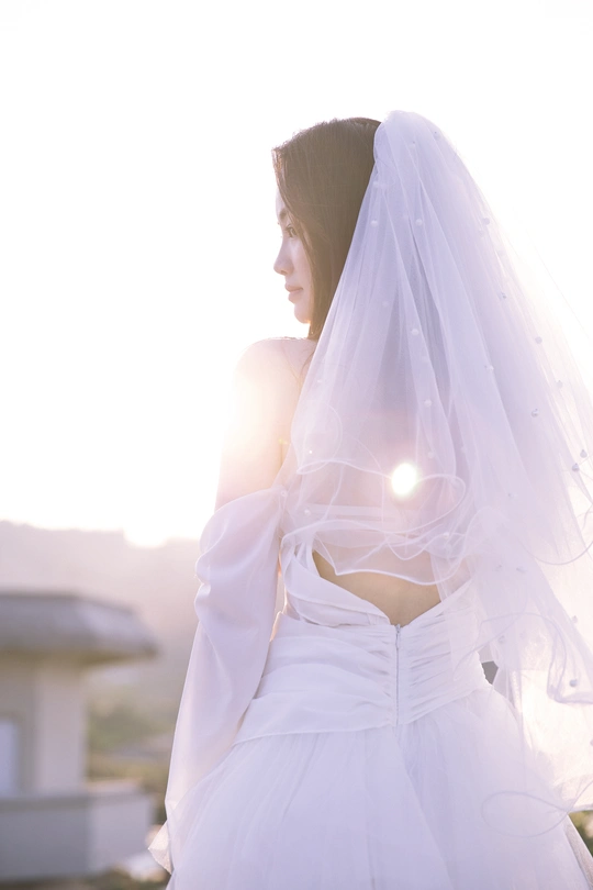 一位美丽的年轻女子穿着一件白色连衣裙和面纱站在屋顶上 太阳在她的身后。