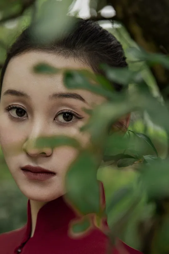 一位穿着红外套的年轻女子在摄影时摆姿势 背景中有绿色的树木和灌木丛。