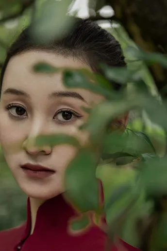 一位穿着红外套的年轻女子在摄影时摆姿势 背景中有绿色的树木和灌木丛。