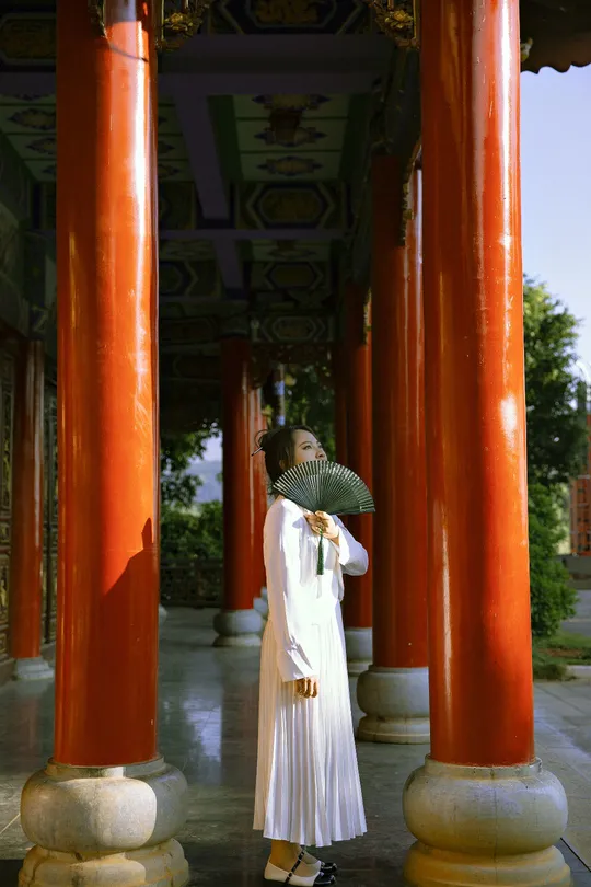 一位穿着白色长袍 手持雨伞 站在寺庙内的红色柱子下的女人