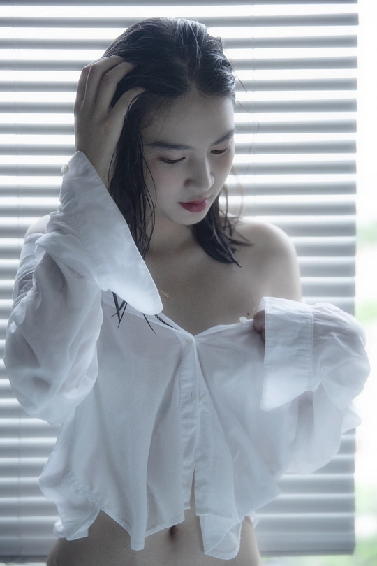 一位年轻女子穿着一件白色衬衫站在窗前 窗帘是关闭的。