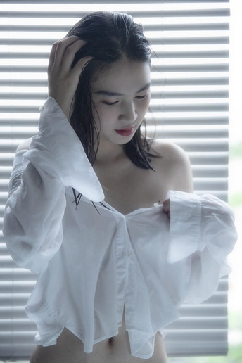 一位年轻女子穿着一件白色衬衫站在窗前 窗帘是关闭的。