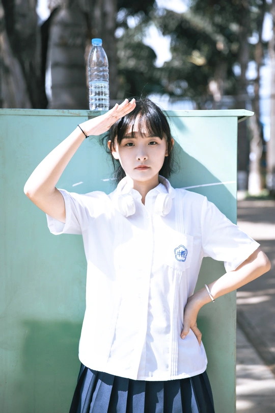 一个穿着高中制服的女孩站在外面 双手抱头 手里拿着一瓶水。