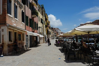 许多人坐在带有遮阳伞的鹅卵石街道上的户外桌子旁。