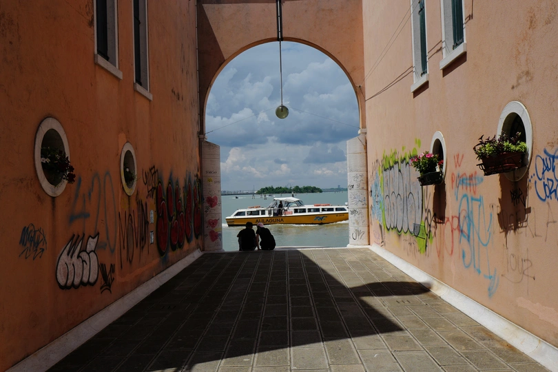 一条街道 墙壁上有涂鸦 街道对面有一个拱门 门内有一艘船在水中。