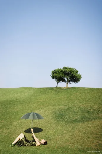 一个女人躺在绿草丛生的小山丘上 顶部有一棵树 而另一个人则在绿草地上放松。