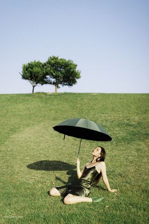 一个穿着黑色连衣裙的女人拿着黑色雨伞坐在草地上 背景中有一棵树。
