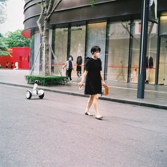 一个穿着黑色连衣裙的女人带着行李箱走在街上 一辆电动滑板车在建筑物前的人行道上行驶。