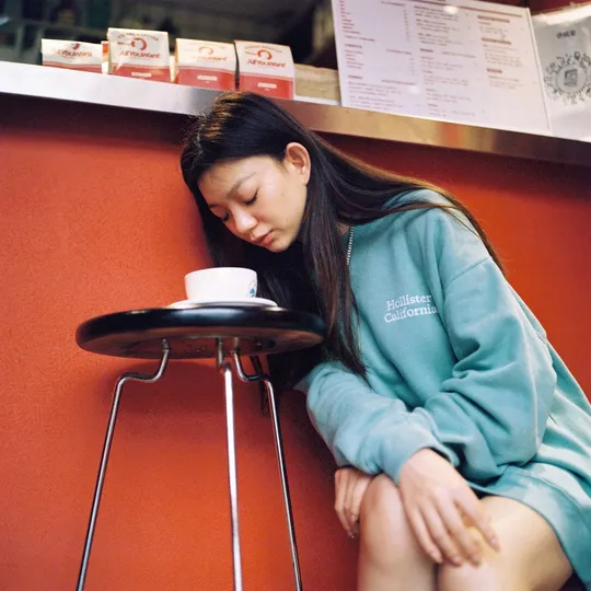 一个年轻女子坐在桌子旁边的一把小凳子上 手里拿着一杯咖啡。