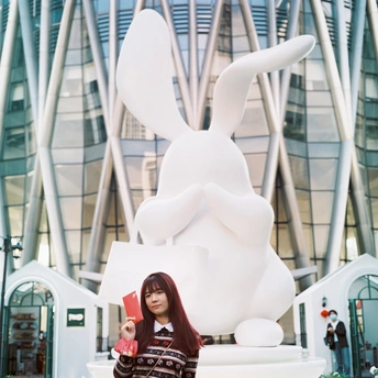 一位年轻女子手持红色书籍站在一栋建筑前的大型白色兔子雕塑前。