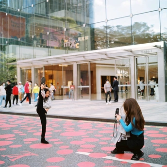 一位年轻女子站在一栋玻璃建筑前 人们正在拍照 而地面上也有照片。