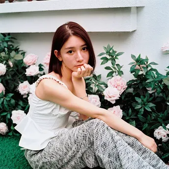 一位穿着白色裤子的年轻女子坐在墙前的草地上 周围有花。