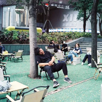 一个男人坐在公园里的一棵树下的一把椅子上。