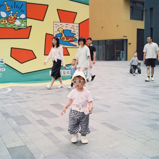 一个小女孩戴着太阳镜和帽子走在街上 而其他人站在背景中。