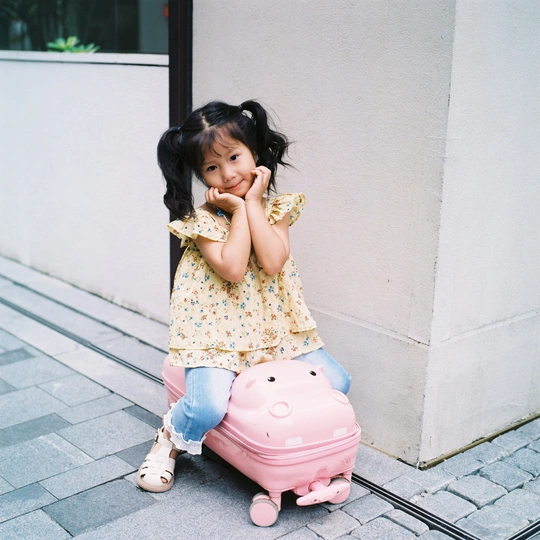 一个小女孩坐在人行道旁边的一个粉色箱子上 旁边有一栋建筑。