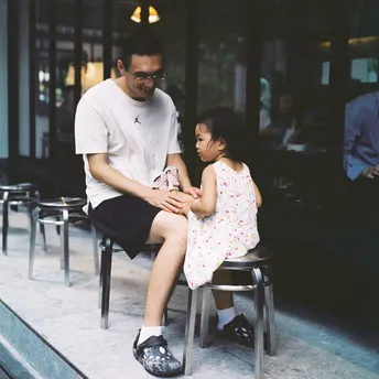 一个男子坐在窗前的一把椅子上 一个小女孩坐在他前面的椅子上。