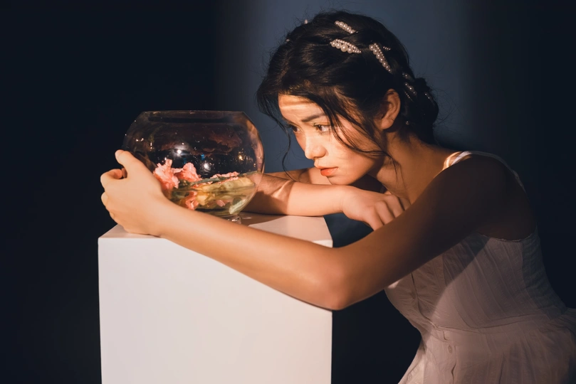一个年轻的女人手里拿着一碗花 看着白色底座上鱼缸里的鱼
