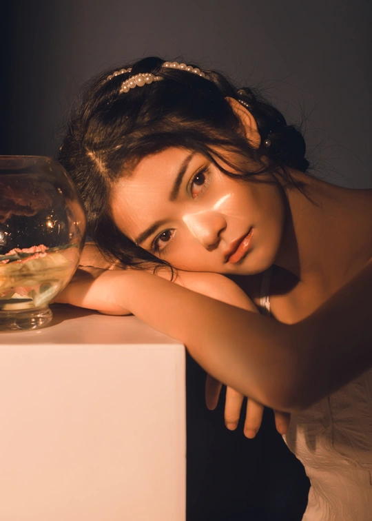 一位年轻女子拿着一条金鱼坐在白色桌子上水族馆旁边的碗里