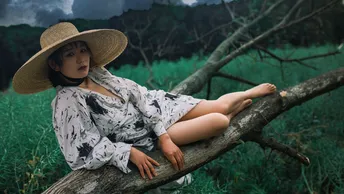 一位穿着连衣裙和帽子坐在树树枝上的年轻女子在田野中