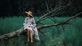 一位穿着连衣裙和帽子坐在树树枝上的年轻女子在田野中