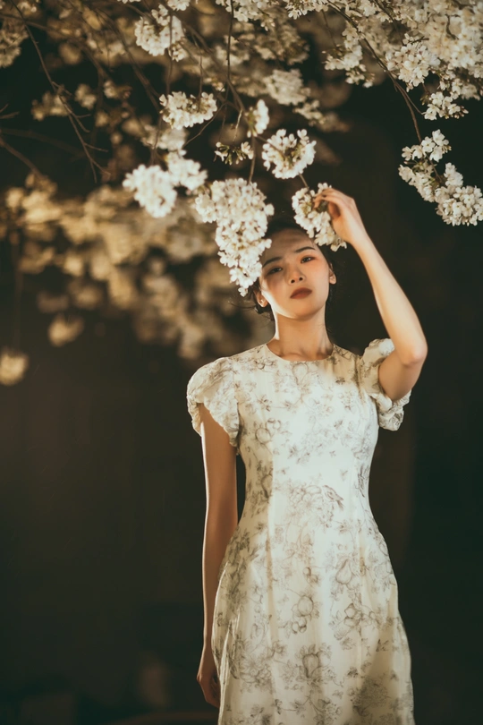 一位穿着白色连衣裙的年轻女子站在一棵点亮的树前 手里拿着花。