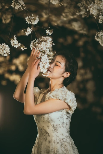 一位穿着白色连衣裙的年轻女子在夜晚闻着树上的白色花朵