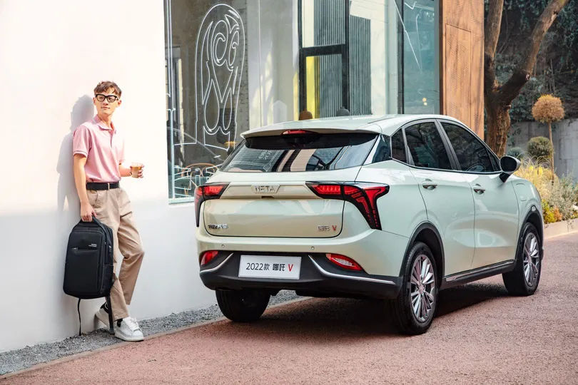 一个年轻男子站在一栋建筑外 旁边停着一辆车 有人靠在墙上 手里拿着一个箱子。