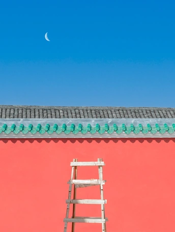 一根倾斜的木材梯子靠在红色墙壁上 背景是蓝色的天空和月亮。