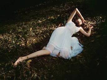 一位穿着白色连衣裙的年轻女子躺在森林中的一棵树附近 在地上。