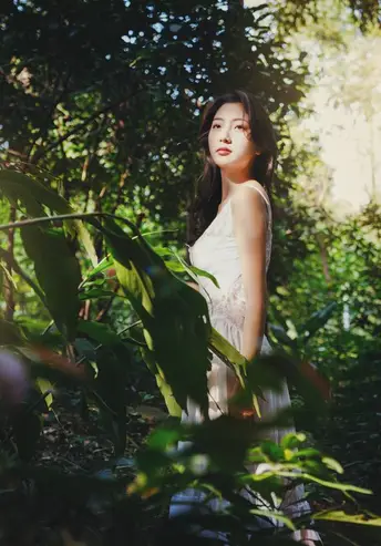 一位穿着白色连衣裙的年轻女子在森林中为肖像摆姿势 太阳照亮了树木。