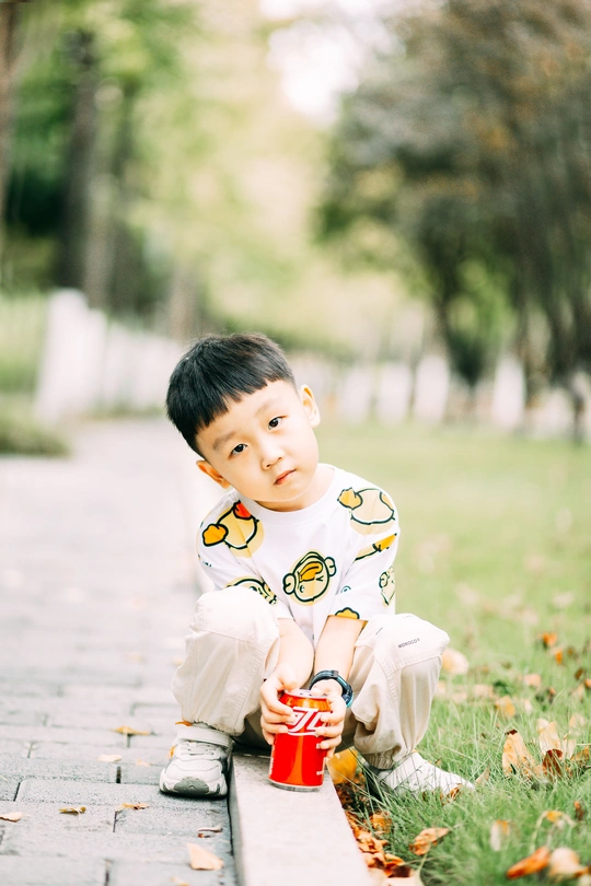 一个穿着睡衣的小男孩坐在公园的人行道上 手里拿着一个红杯子和一罐苏打水放在草地上